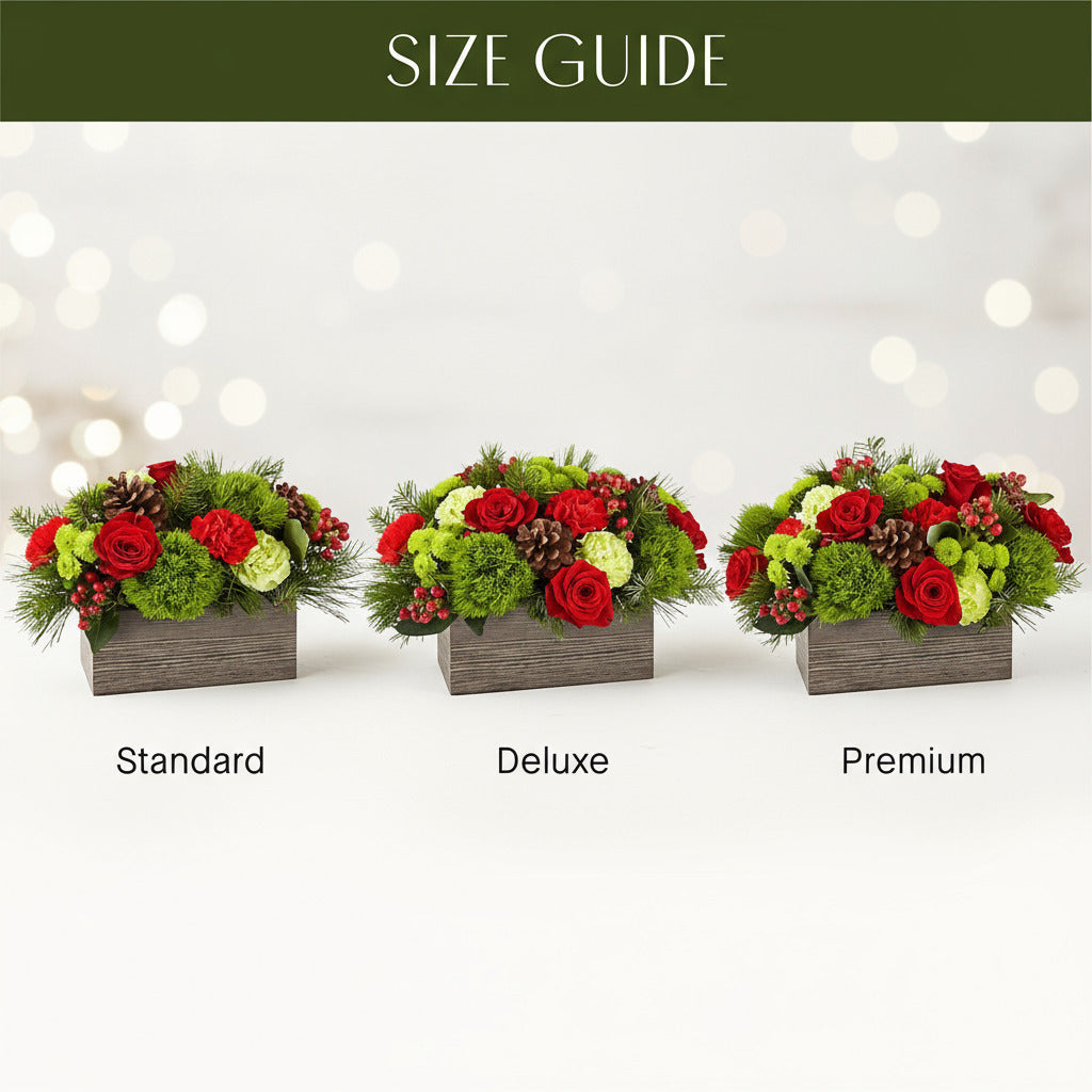 A floral centerpiece arrangement featuring a variety of holiday-themed botanicals including green balls, carnations, roses, cushion poms and hypericum, with pine cones and a plaid ribbon, displayed in a rectangular box.