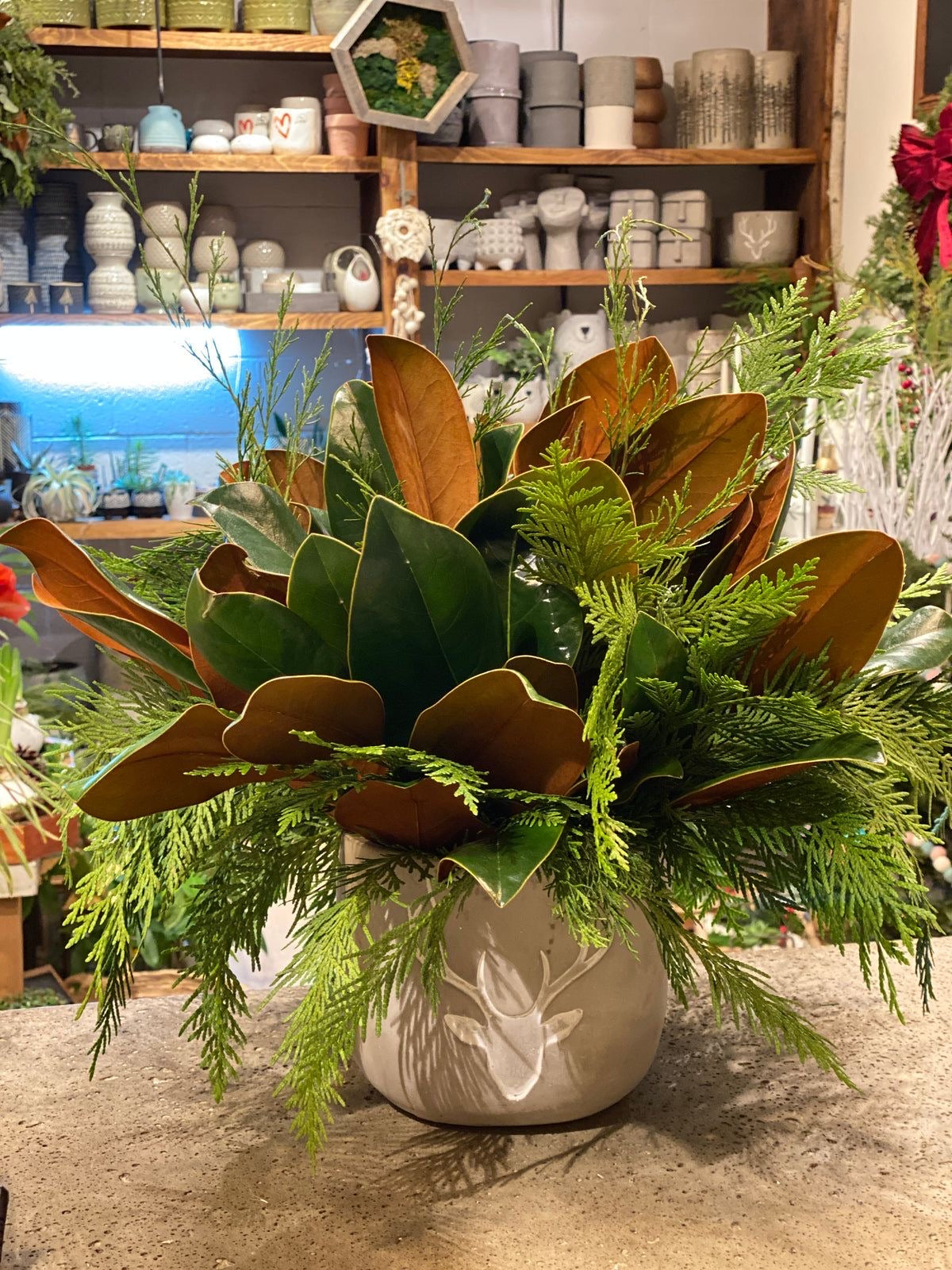 A floral arrangement with green leaves and brown tones, displayed in a white vase, set against a shelf with various pottery items in the background. Magnolia Dream bouquet with magnolia leaves and greenery arranged in a low vase, perfect for cozy fall and holiday decor
