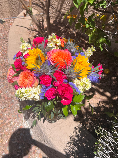 Colorful bouquet with hot pink, violet, yellow, lavender, and peach blooms arranged with seasonal greenery.
