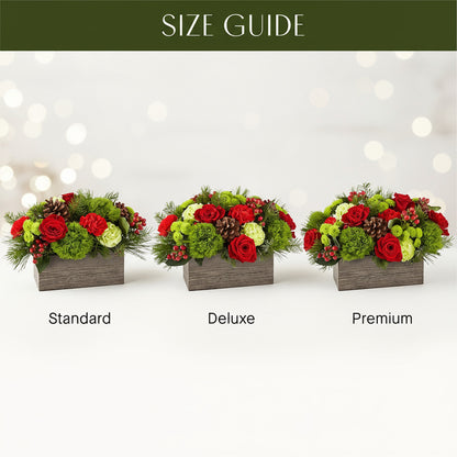A floral centerpiece arrangement featuring a variety of holiday-themed botanicals including green balls, carnations, roses, cushion poms and hypericum, with pine cones and a plaid ribbon, displayed in a rectangular box.