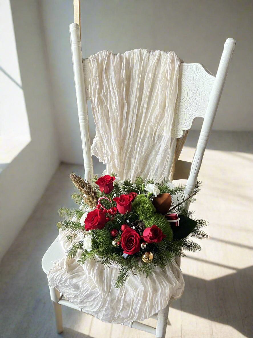 A tabletop centerpiece featuring a red container with a mix of green and red decorations including pinecones, berries, and artificial apples, arranged on a white chair with a lacey white fabric.
