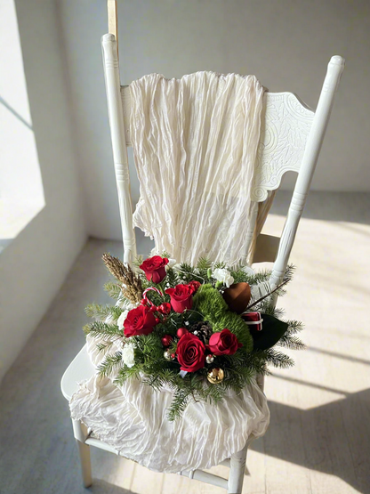 A tabletop centerpiece featuring a red container with a mix of green and red decorations including pinecones, berries, and artificial apples, arranged on a white chair with a lacey white fabric.