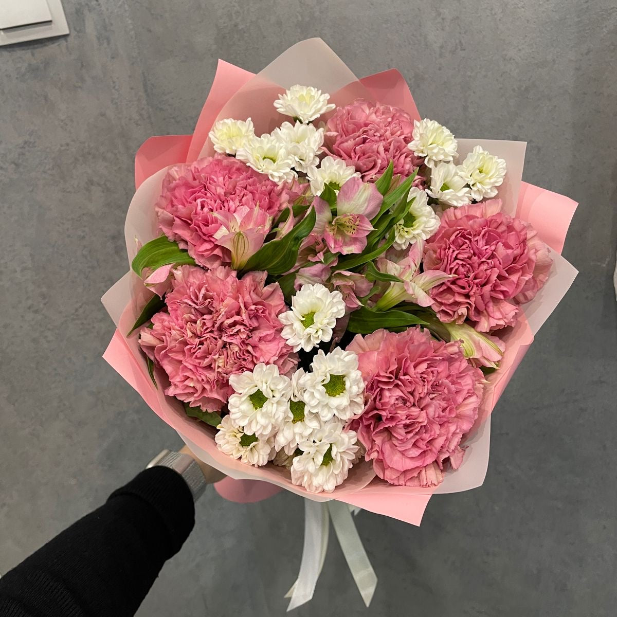 A bouquet of flowers featuring carnations and roses in shades of pink, white, and green, wrapped in pink paper.
