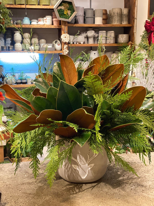A floral arrangement with green leaves and brown tones, displayed in a white vase, set against a shelf with various pottery items in the background. Magnolia Dream bouquet with magnolia leaves and greenery arranged in a low vase, perfect for cozy fall and holiday decor